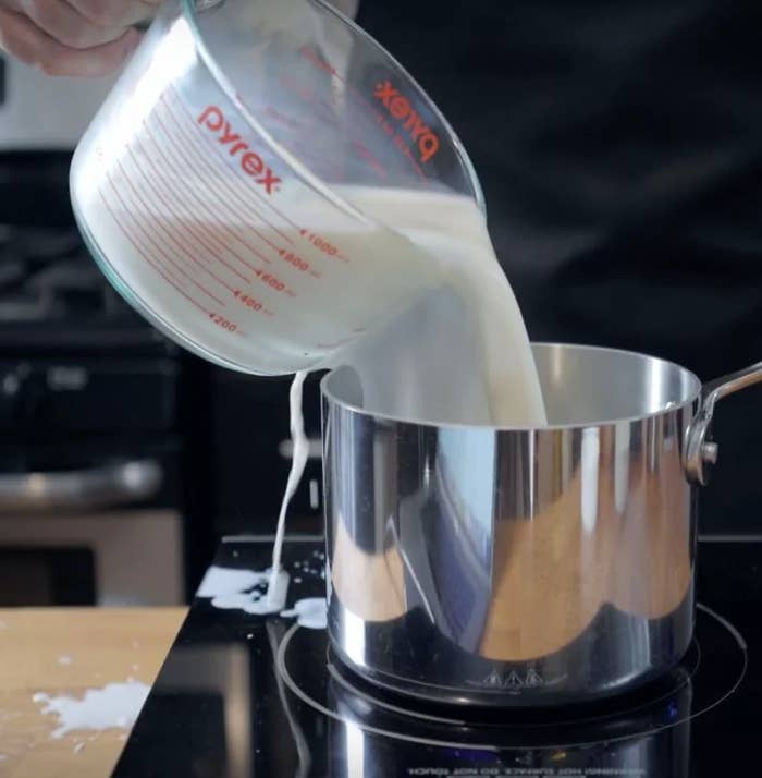 Measuring cup pouring milk into a metal saucepan on a stovetop, with some milk spilling over the edge