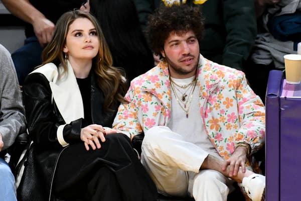 Two people sitting courtside at a sports event. One wears a black and white coat, the other a floral jacket with layered necklaces