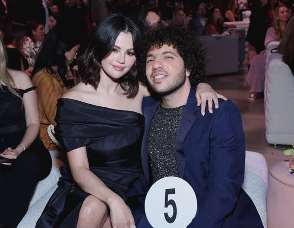 Two people sitting; one holds a number 5 paddle. The person on the left wears a stylish off-shoulder dress. They are at an event with seated guests