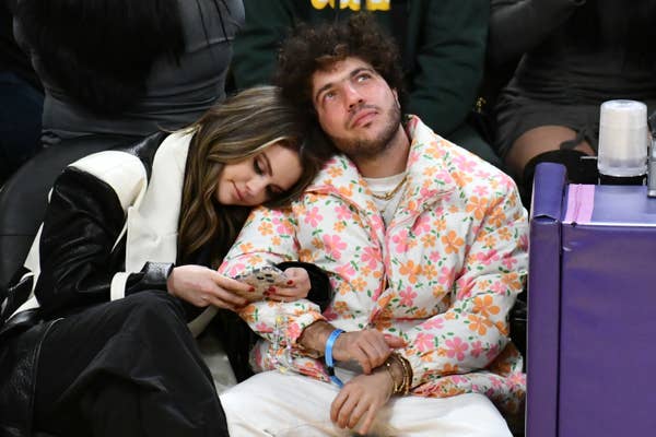 Two people sitting courtside at a sports event. One person, in a stylish floral jacket, looks up while the other rests their head on their shoulder