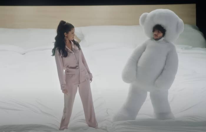 Woman in silk pajamas stands facing a person in a large fluffy teddy bear costume, on an oversized bed