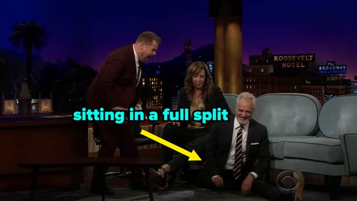 Talk show host and guests laugh as a man in a suit performs a split on the studio floor during a lively late-night segment