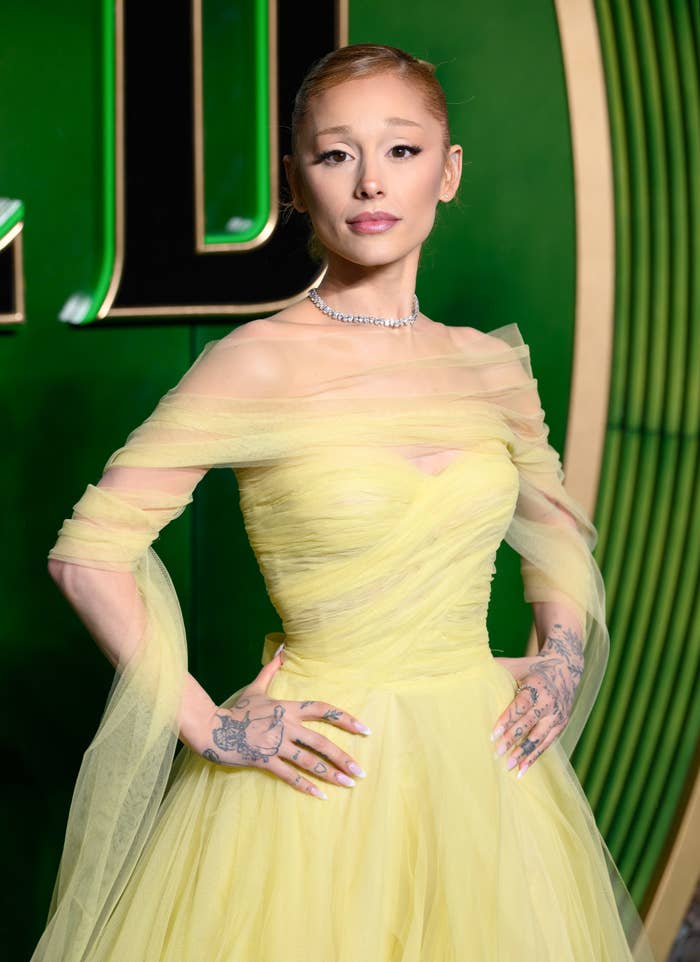 Person posing on a red carpet wearing an elegant off-shoulder gown with sheer layers, adorned with tattoos and a necklace