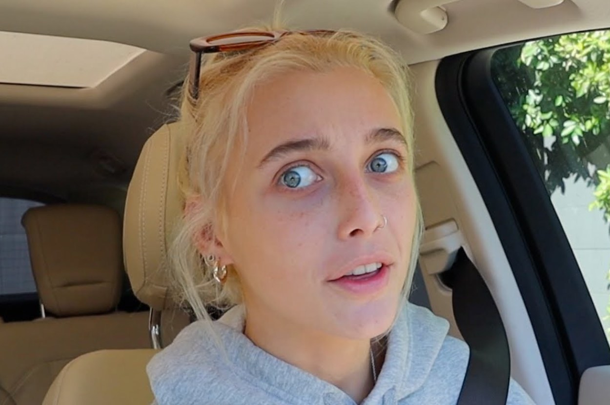 Person with sunglasses on head, seated in a car, looking towards the camera with a neutral expression. Trees visible through the window