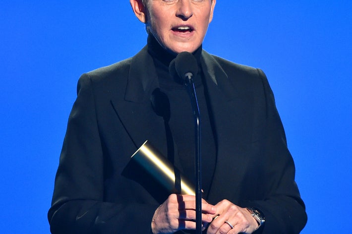 Ellen DeGeneres in a suit holding an award, speaking on stage at an event