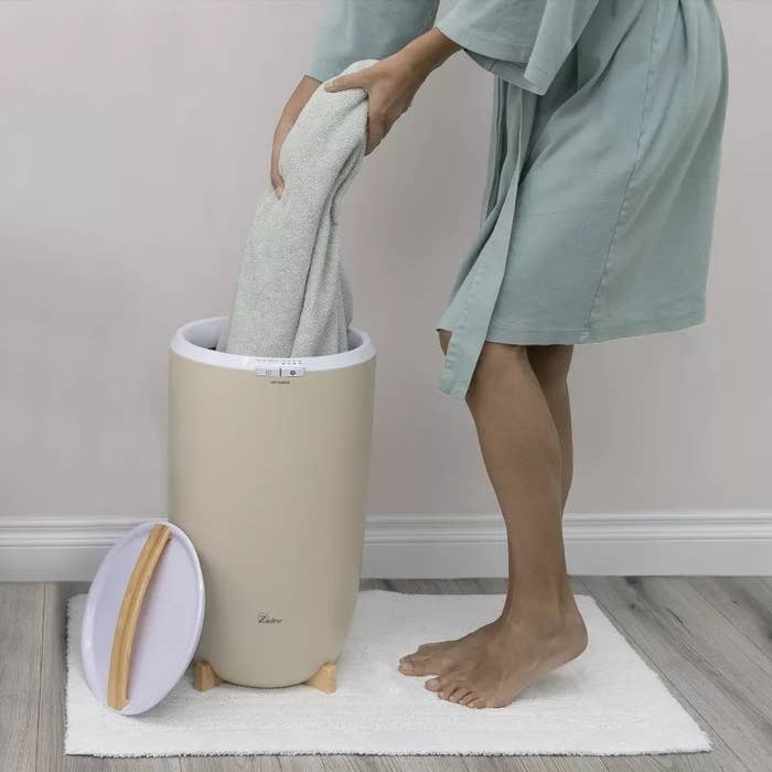 Person placing a towel into a beige laundry hamper with a lid, standing on a white rug in a minimalist room