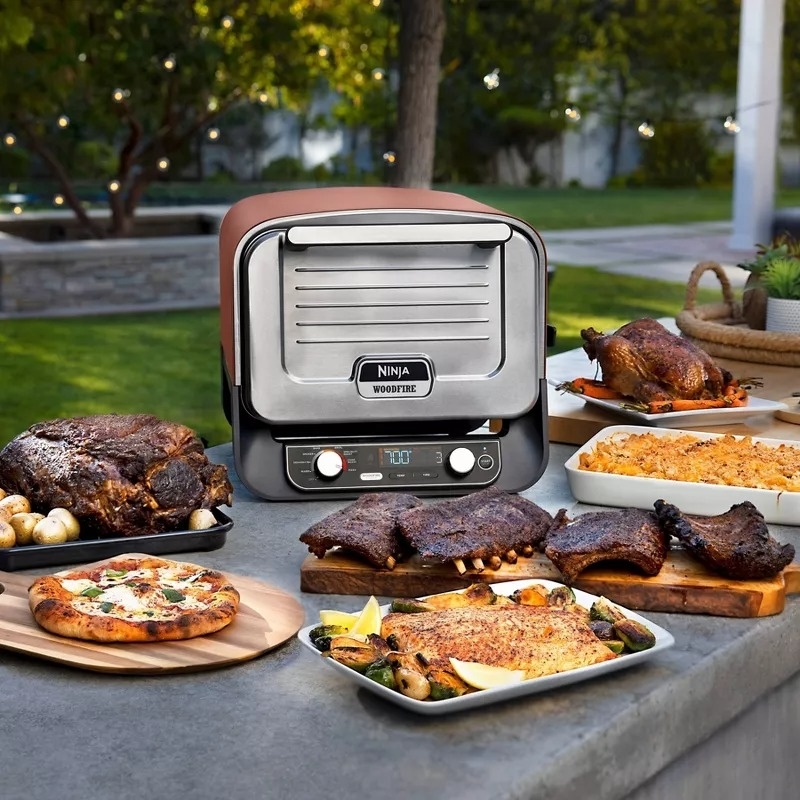 Outdoor kitchen scene with a Ninja Woodfire grill showcasing a variety of cooked meats and dishes