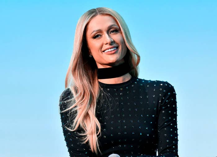 Woman in elegant, studded black outfit smiles while posing, with sky in the background