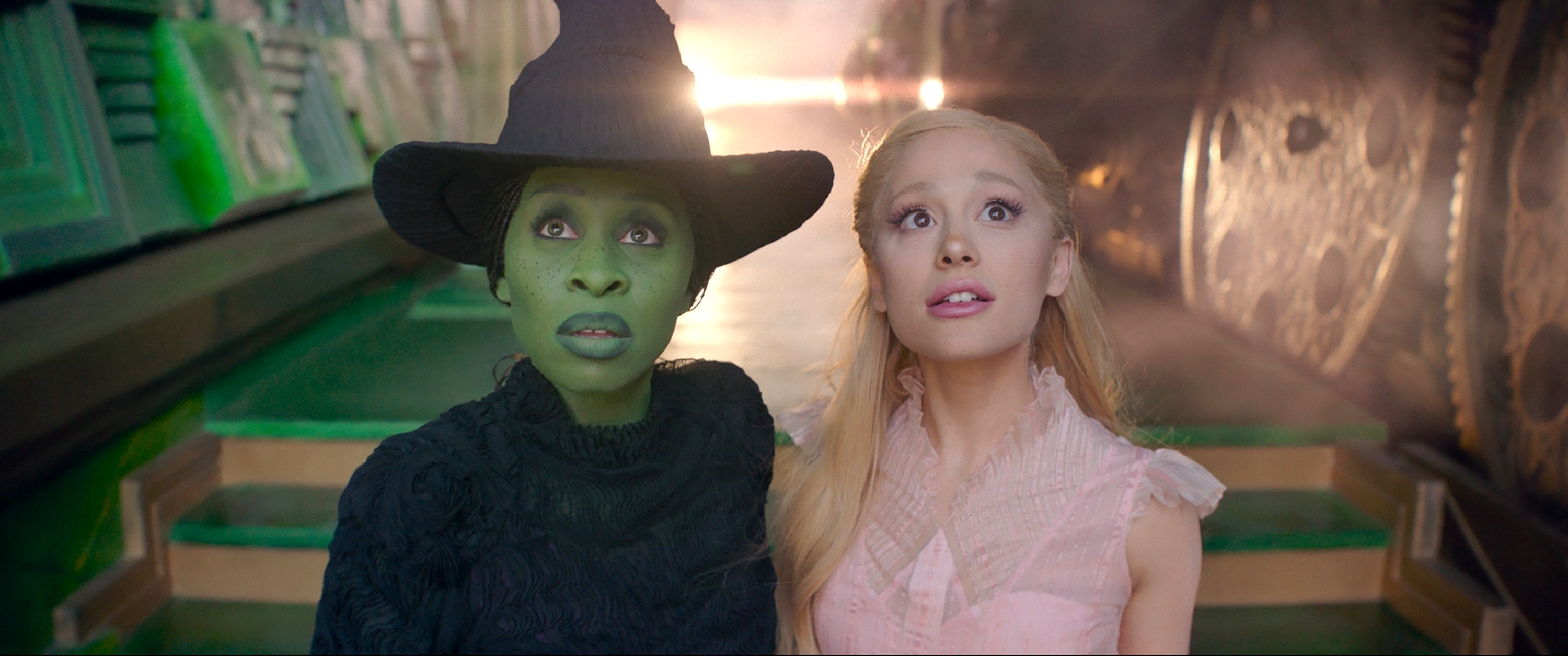 Cynthia Erivo and Ariana Grande in fantasy costumes: one with a witch hat and green face, wearing a dark ensemble; the other in a light, sleeveless dress, both gazing upward