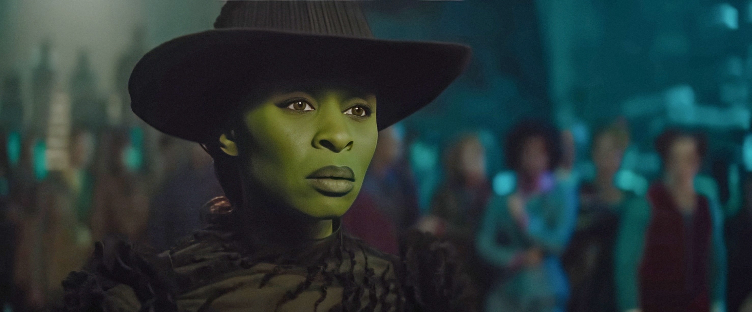 Cynthia Erivo in a black hat and textured top with green face paint, looking intently forward in a dramatic setting