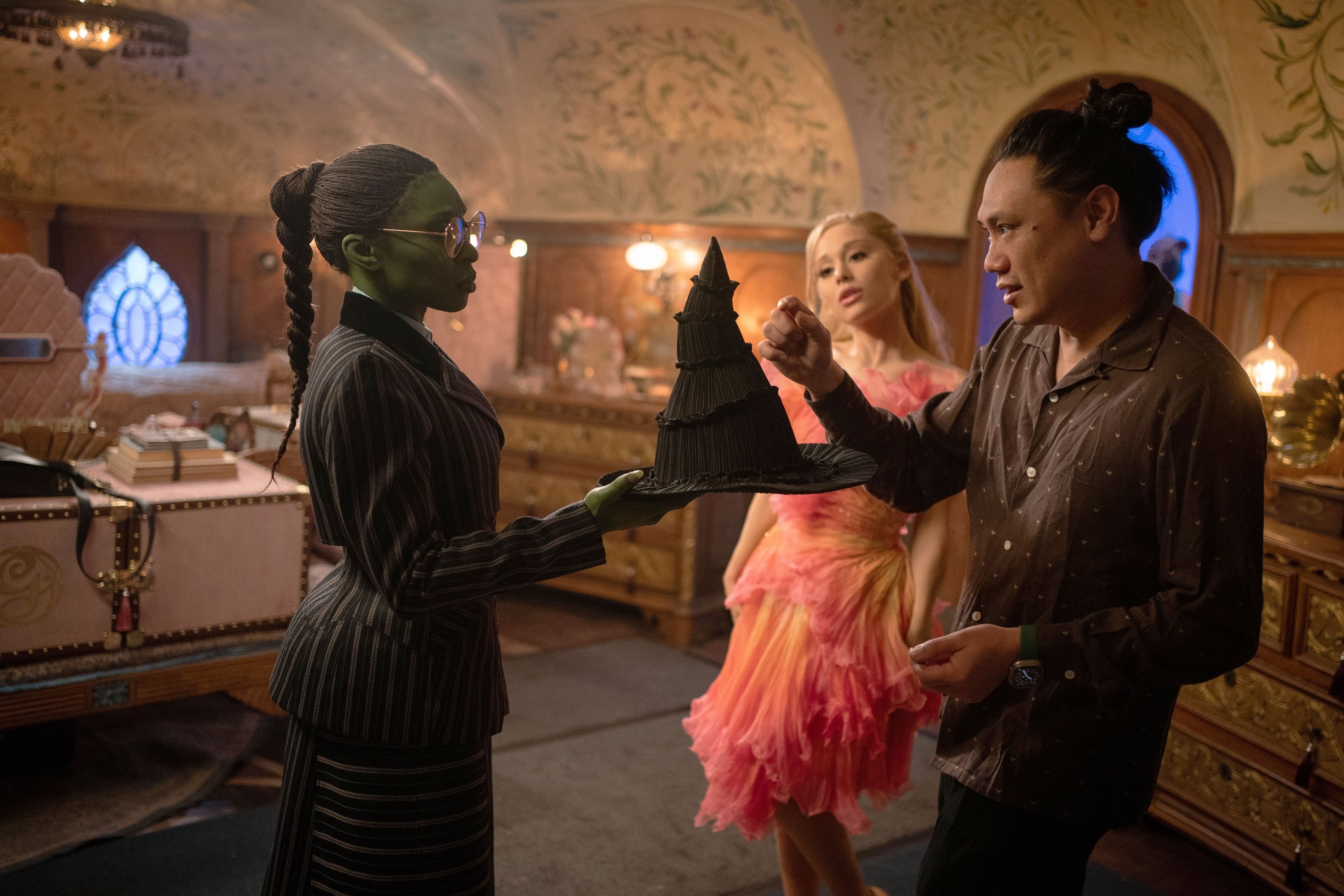 Cynthia Erivo, Ariana Grande, and Jon M. Chu in a ornately decorated room, one holds a pointed black hat. Another wears a frilly dress