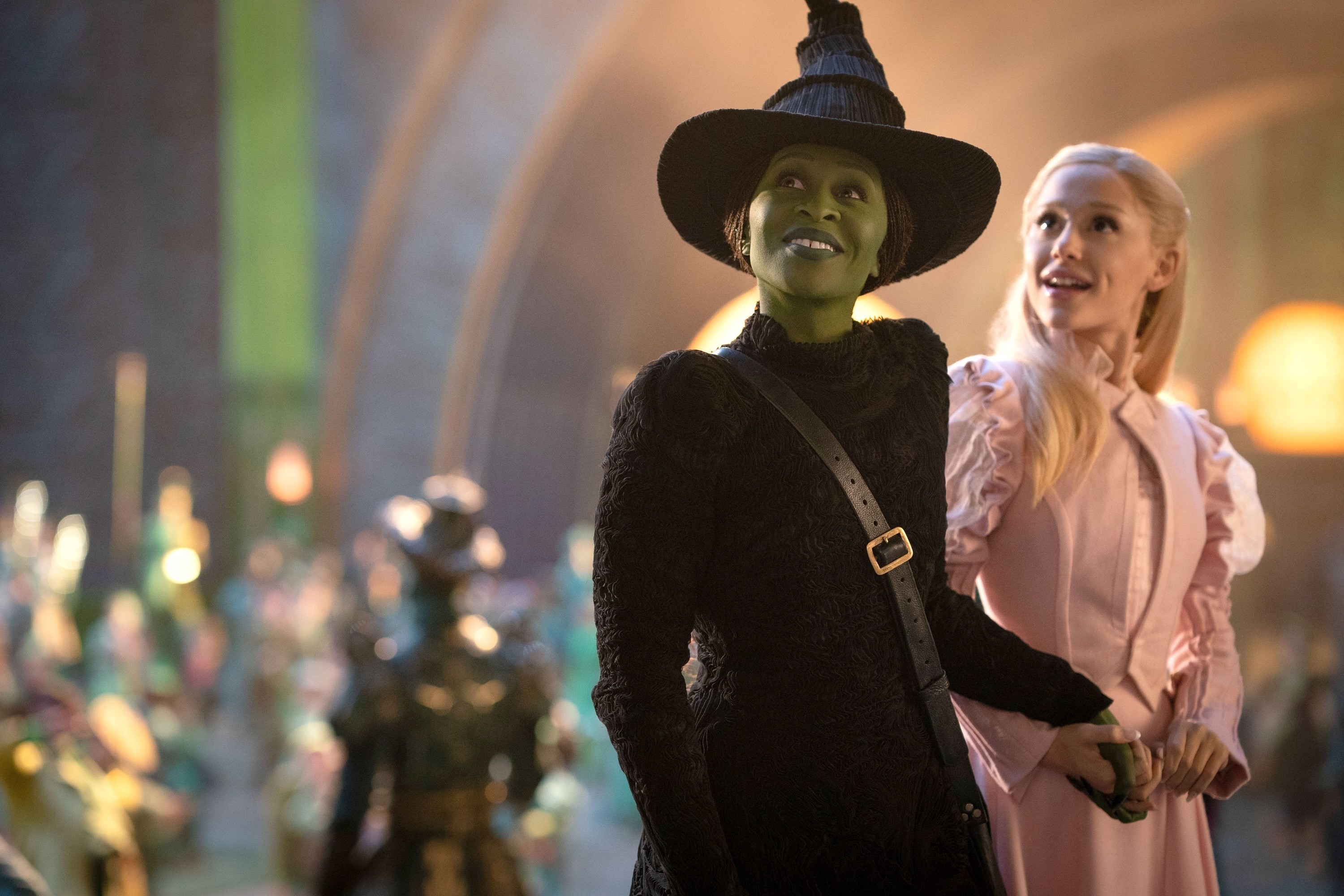 Cynthia Erivo and Ariana Grande in theatrical costumes, one in a witch outfit with a pointed hat, appear in a whimsical setting, holding hands and smiling