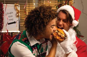 Two people in festive clothing, one in a Christmas sweater and the other in a Santa suit, share a sandwich at a holiday-themed photo shoot