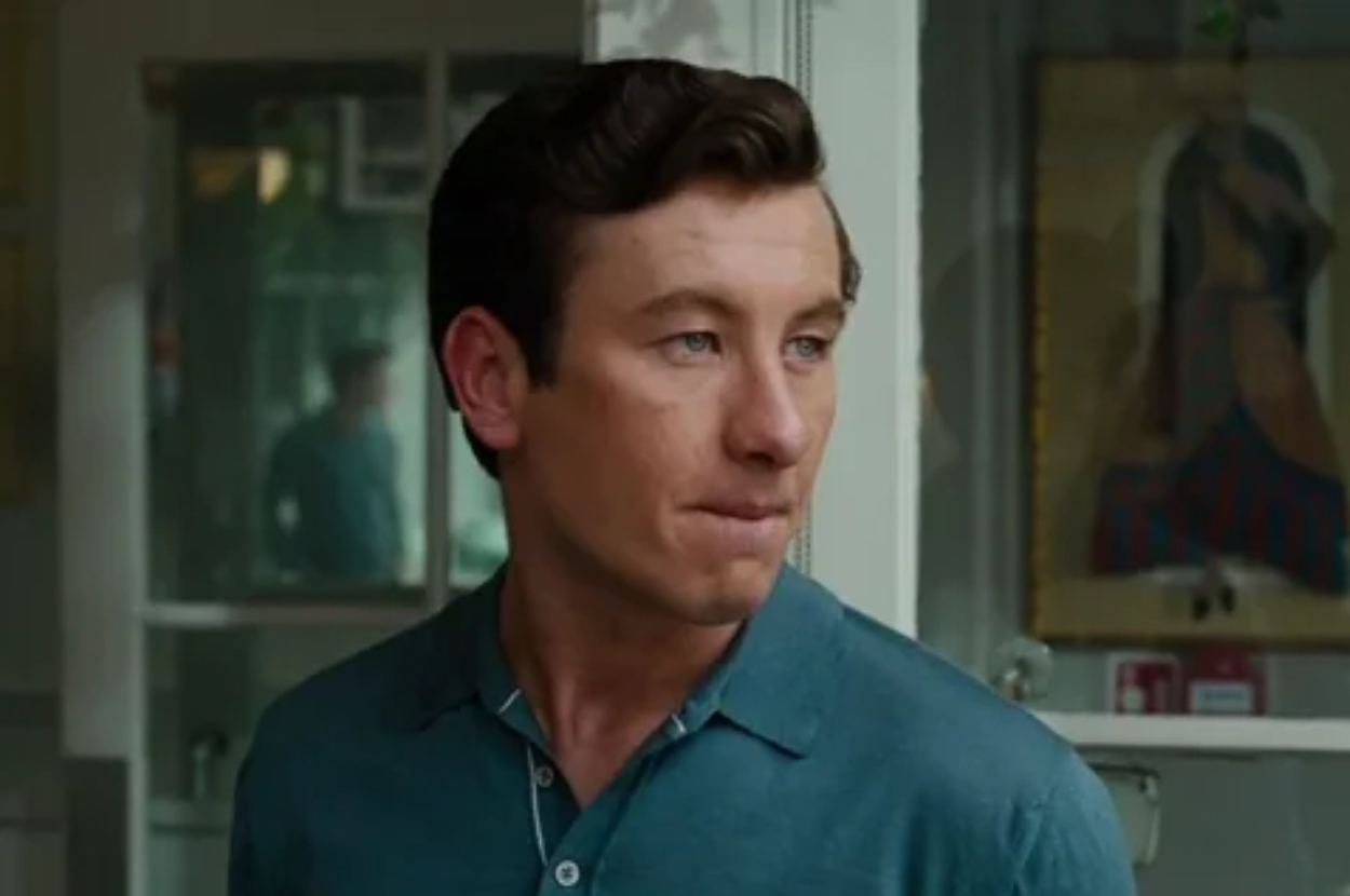 A man with short hair looks to the side, wearing a collared shirt in what appears to be an indoor setting with a framed picture in the background