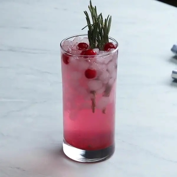 A tall glass with a pink iced drink, garnished with cranberries and a sprig of rosemary, on a light surface