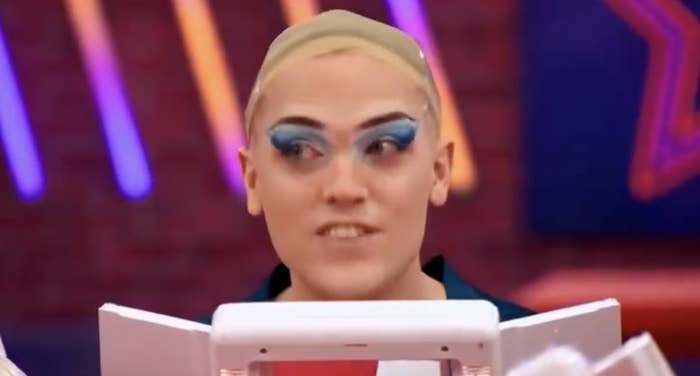 Person with bold eye makeup in a dressing room setting, looking slightly off-camera. Neon lights are blurred in the background