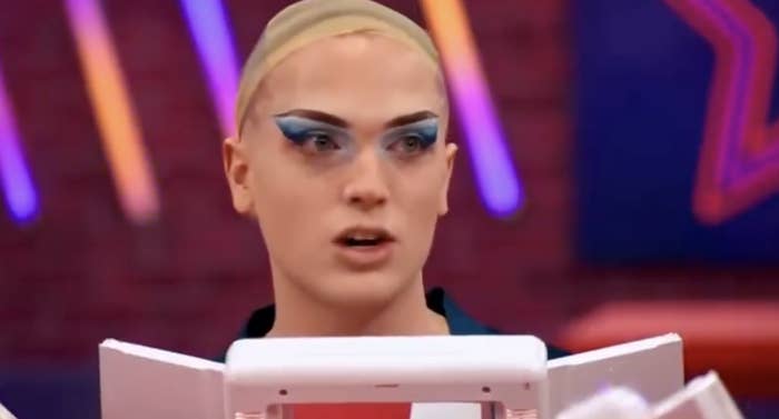 Person with bold makeup and dramatic blue eyeshadow speaking, facing a mirror, in a brightly lit setting with colorful designs in the background