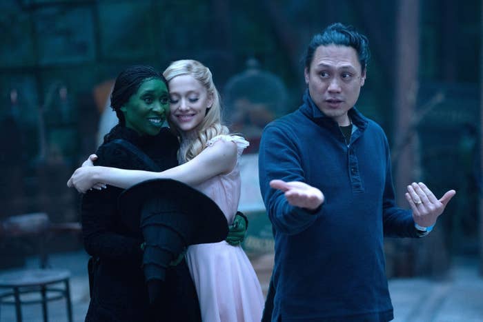 A behind-the-scenes photo of Cynthia Erivo and Ariana Grande hugging as Jon M Chu talks on the set of