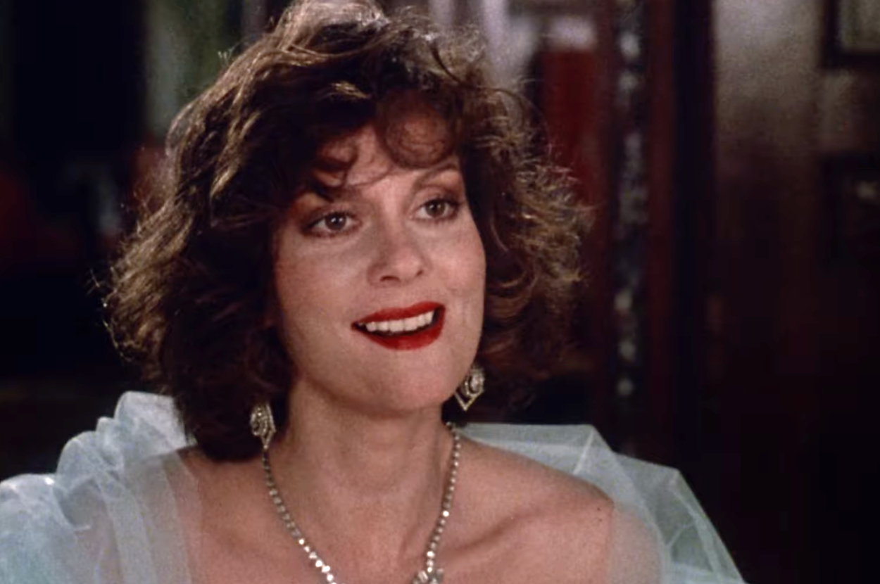 Person smiling, wearing a sheer outfit with earrings and a necklace, in an indoor setting