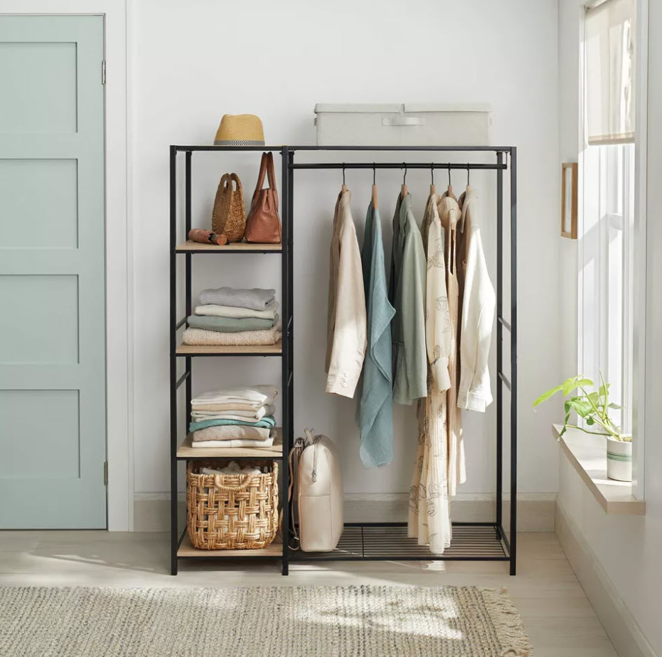 Open closet with neatly hung light-colored clothes, a rattan basket, folded towels, and a bag on shelves next to a sunny window