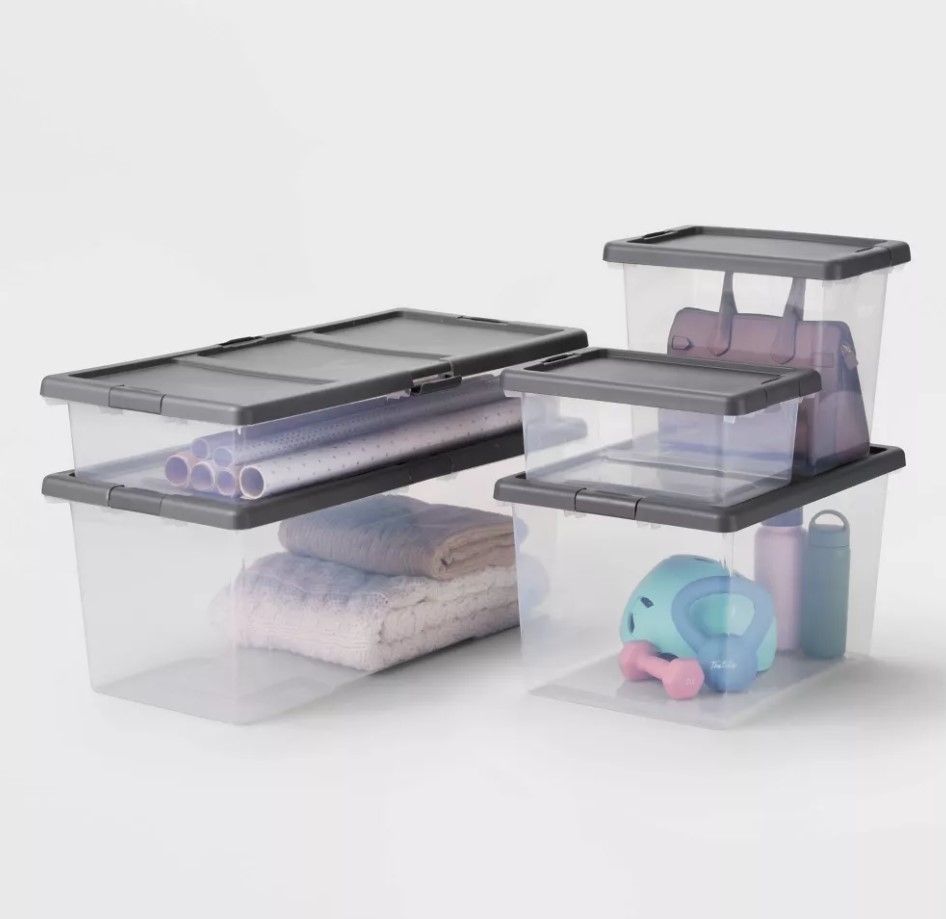 Plastic storage containers filled with various household items, including rolled towels, toys, and a yoga mat, stack neatly on a surface