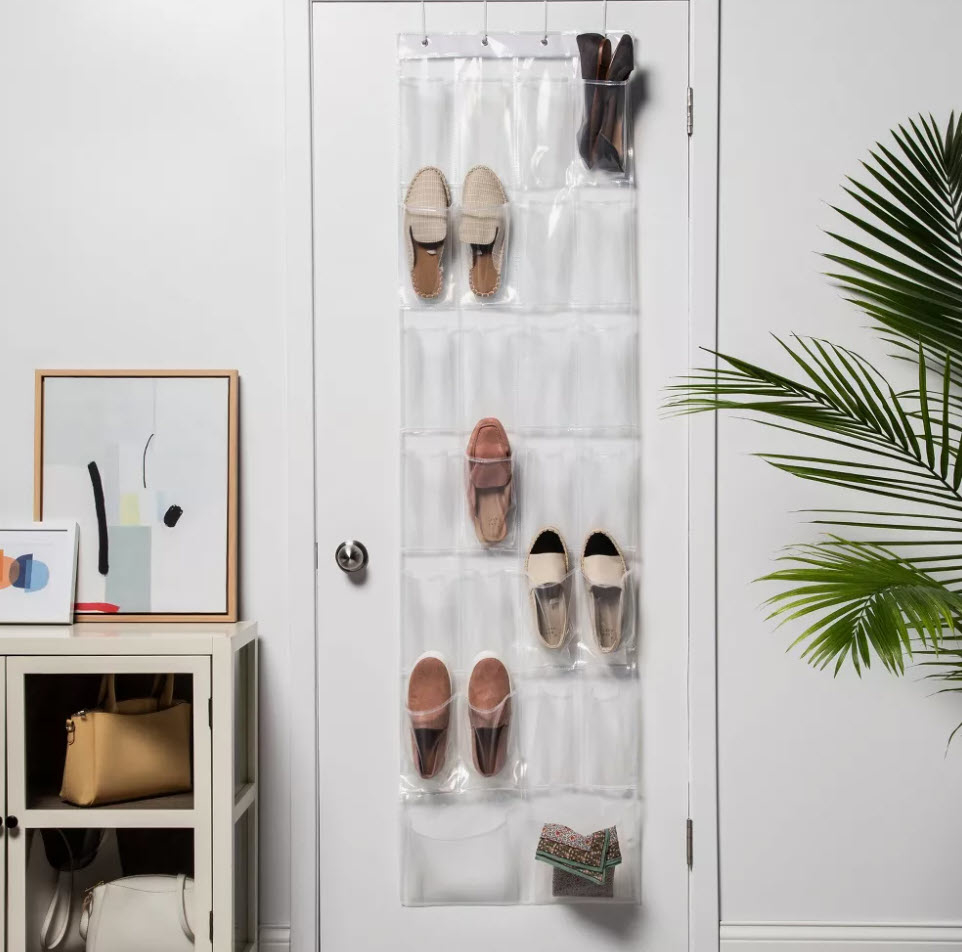 Over-the-door shoe organizer with several shoes and a purse, next to a small cabinet and a framed abstract artwork. A plant is visible in the corner