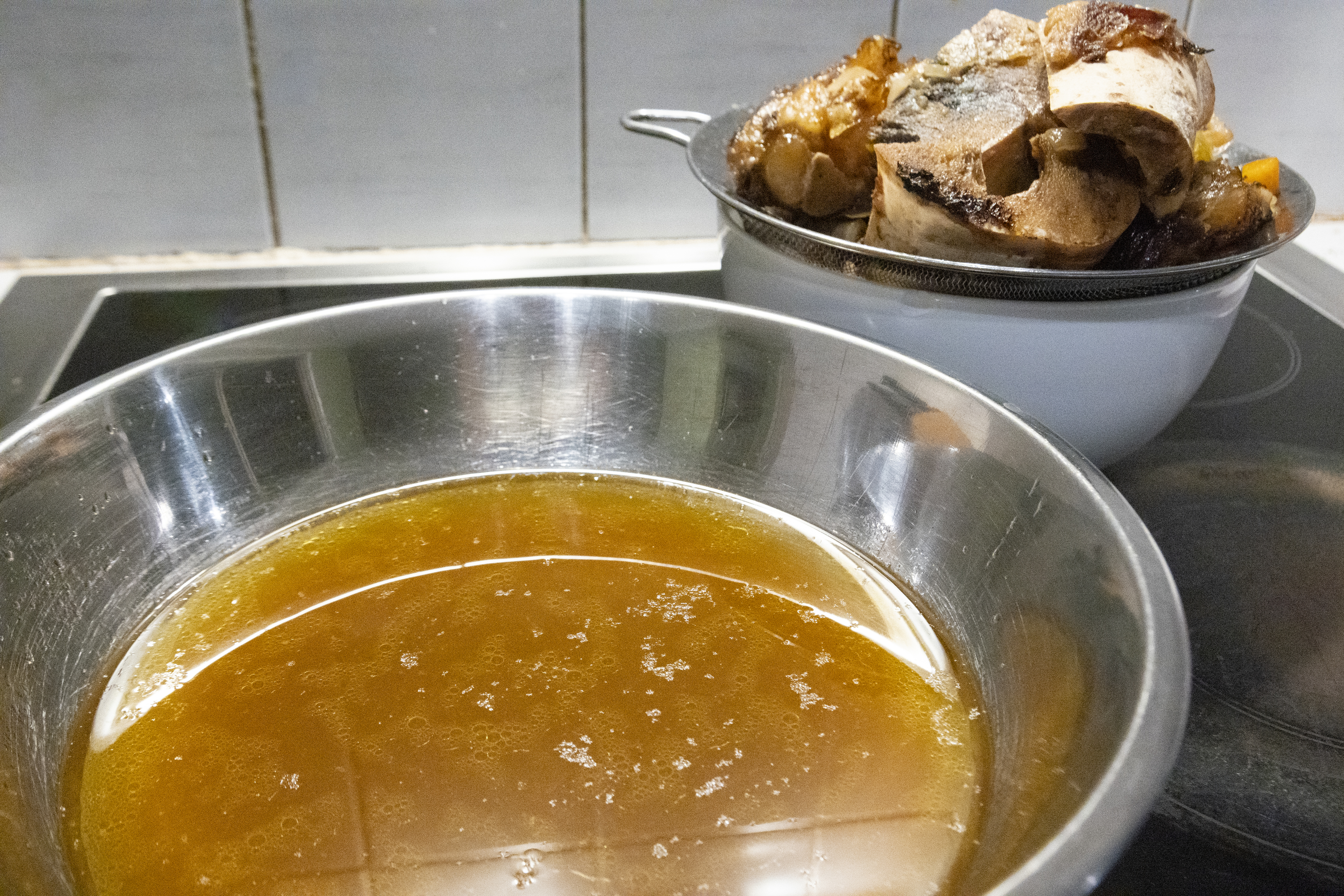 A metal bowl of homemade broth with cooked meat and vegetables in a strainer in the background