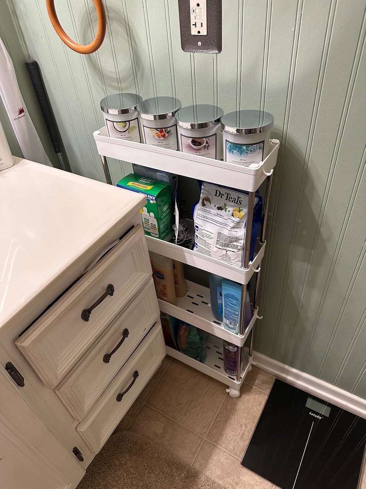 another reviewer's white cart on wheels holding toiletries and household items, placed between a bathroom sink and wall