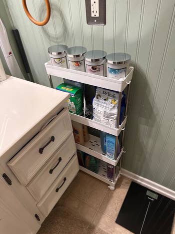 another reviewer's white cart on wheels holding toiletries and household items, placed between a bathroom sink and wall