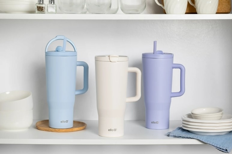 Three insulated travel mugs with handles are displayed on a shelf, featuring different lid styles for easy drinking