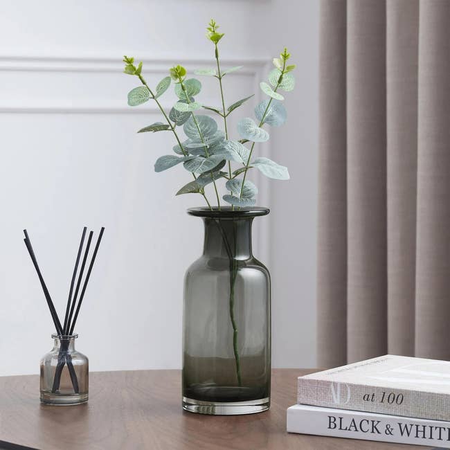 A smoked glass vase with greenery arranged in it, sitting on a wood surface with a reed diffuser and two stacked books
