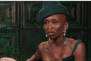 Person wearing a unique hat and elegant jewelry, seated against a textured wall, looking engaged and expressive