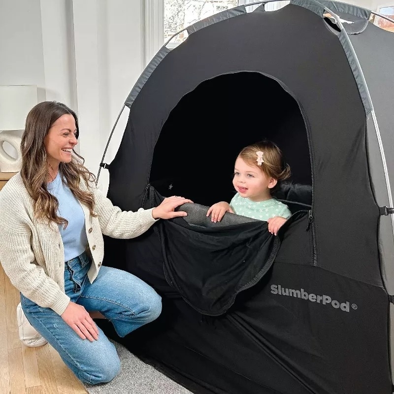 Adult kneels beside child in a SlumberPod indoor privacy tent, smiling. Child peeks out from inside the tent