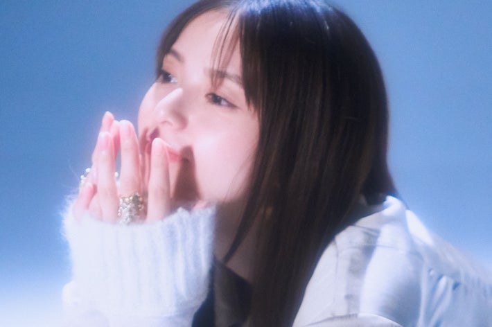 Person with long hair and white gloves kneels and looks thoughtfully upward, wearing a stylish jacket in an artistic setting