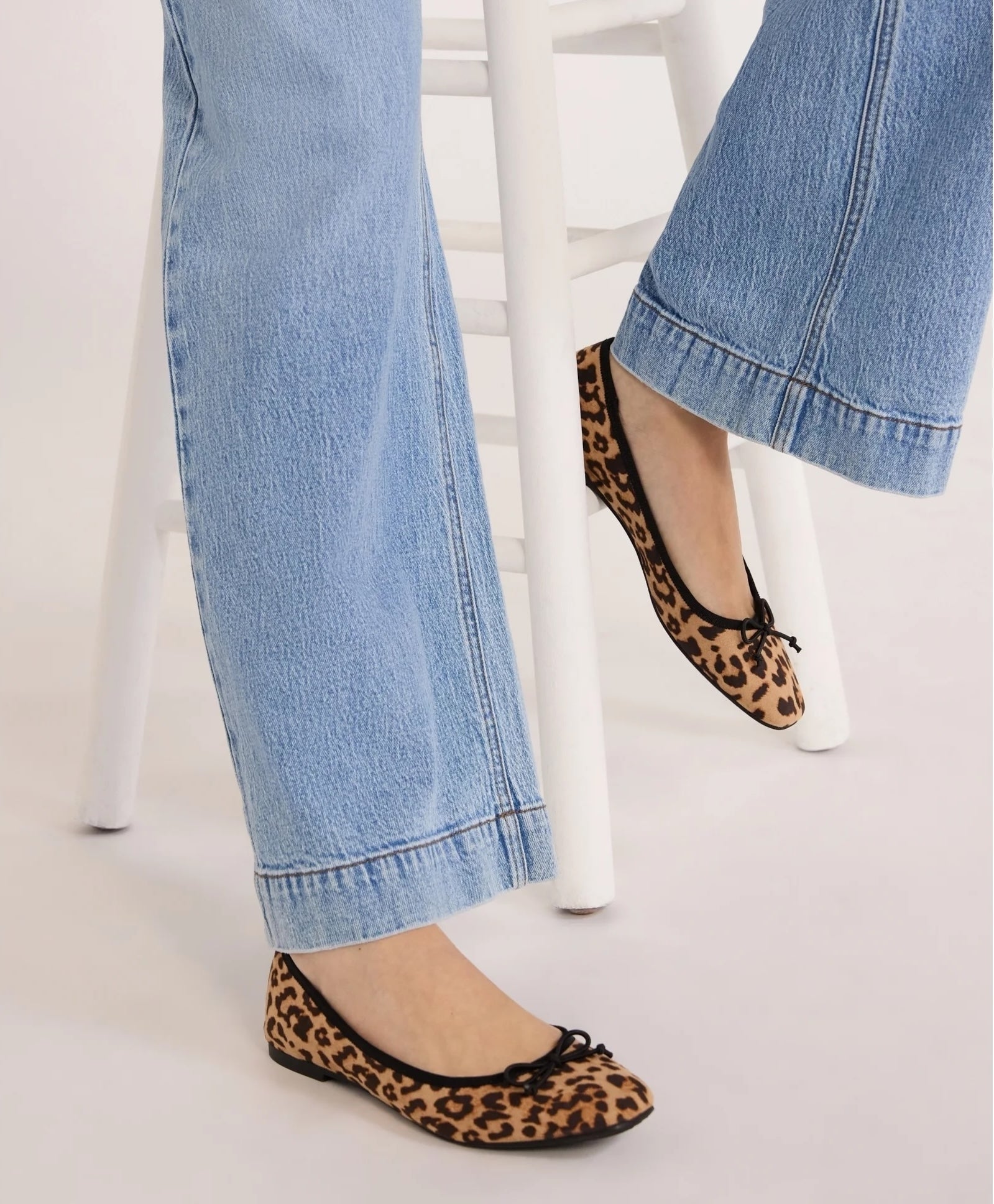 Person wearing blue jeans and leopard print ballet flats, standing by a white stool. Featured in a shopping article