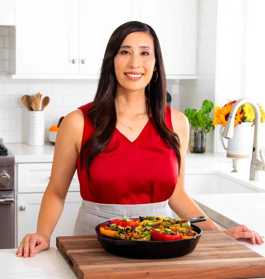 Jessica Gavin, in a kitchen, smiles at the camera, wearing a sleeveless top. They stand behind a tray of colorful, stuffed bell peppers on a cutting board
