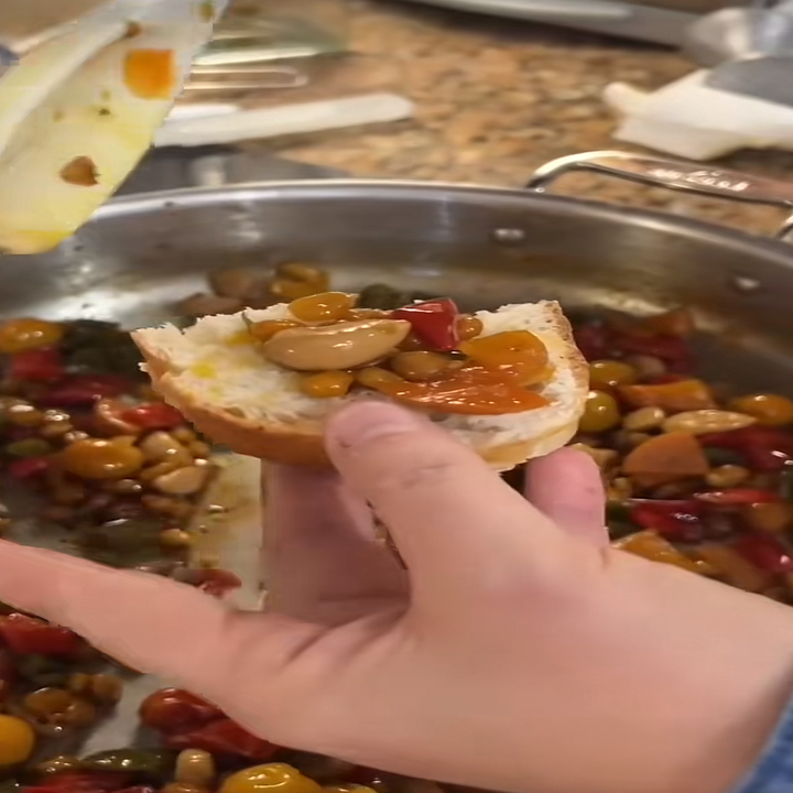 Hand holding a piece of bread with sautéed vegetables and mushrooms from a skillet in the background