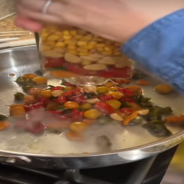 Vegetables and beans are being added to a skillet on a stovetop, creating a colorful medley of ingredients in a kitchen scene