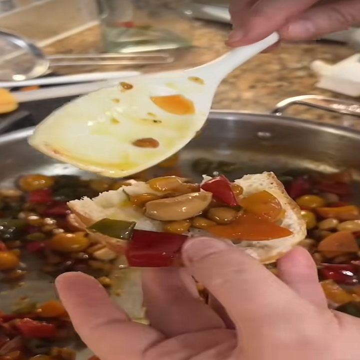 Person spreading a vegetable medley onto a slice of bread with a spoon in a kitchen setting