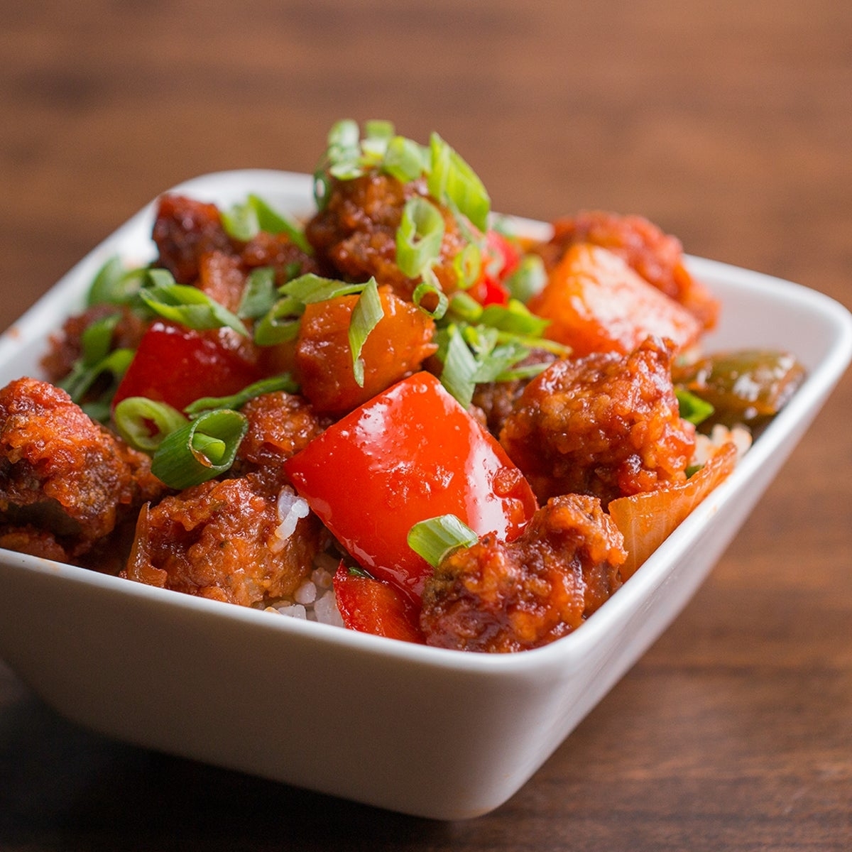 A white square bowl filled with spicy-looking meat, red bell peppers, green onions, and rice, placed on a wooden surface