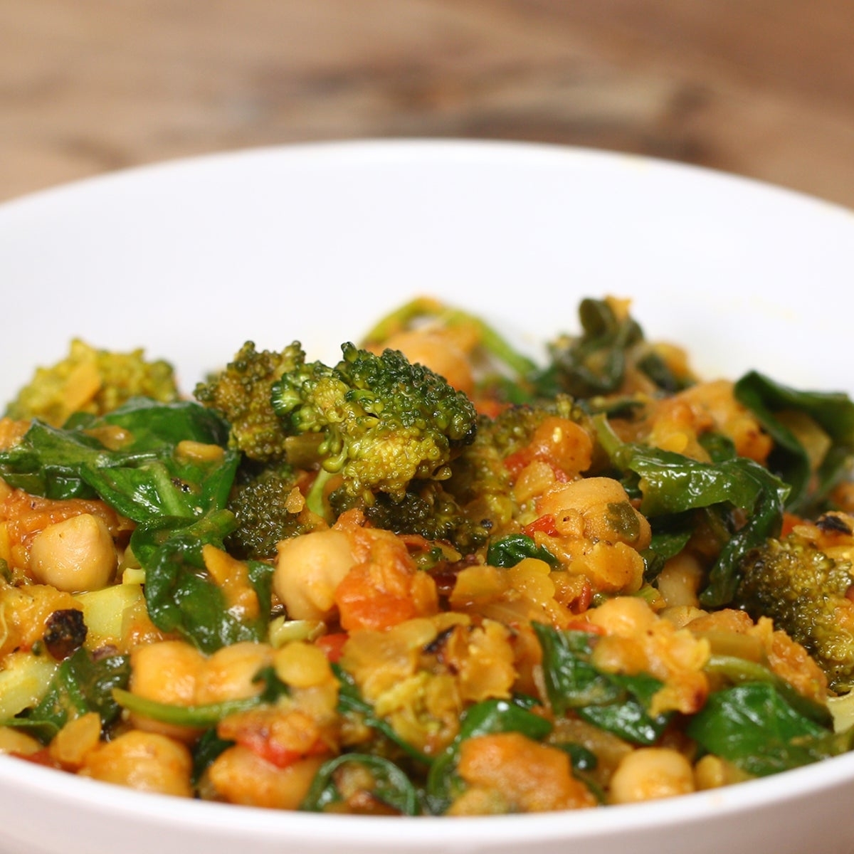 A white bowl filled with a mixed vegetable curry, featuring chickpeas, broccoli, spinach, and tomatoes