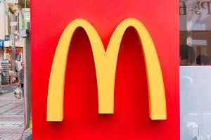マクドナルドの黄色いM字看板が赤い背景に掲示されている。歩道に面した街の景色が一部写っている。