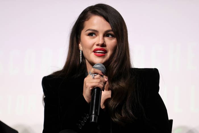 Selena Gomez speaking into a microphone, wearing a blazer and long earrings