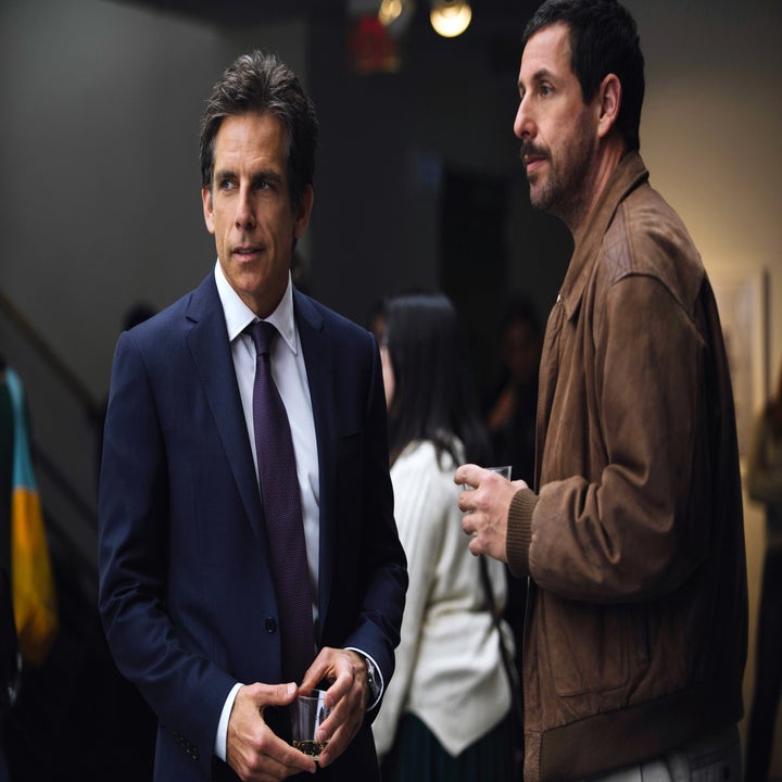 Two men conversing, one in a suit and tie, and the other in a casual jacket holding a drink. They are in a dimly lit room with other people in the background