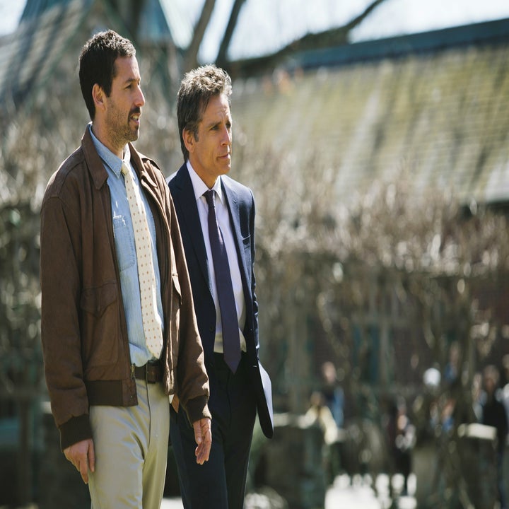Two men in suits walking outside together, engaged in conversation. One wears a tie and a jacket, the other a suit and no tie. Trees and buildings in background