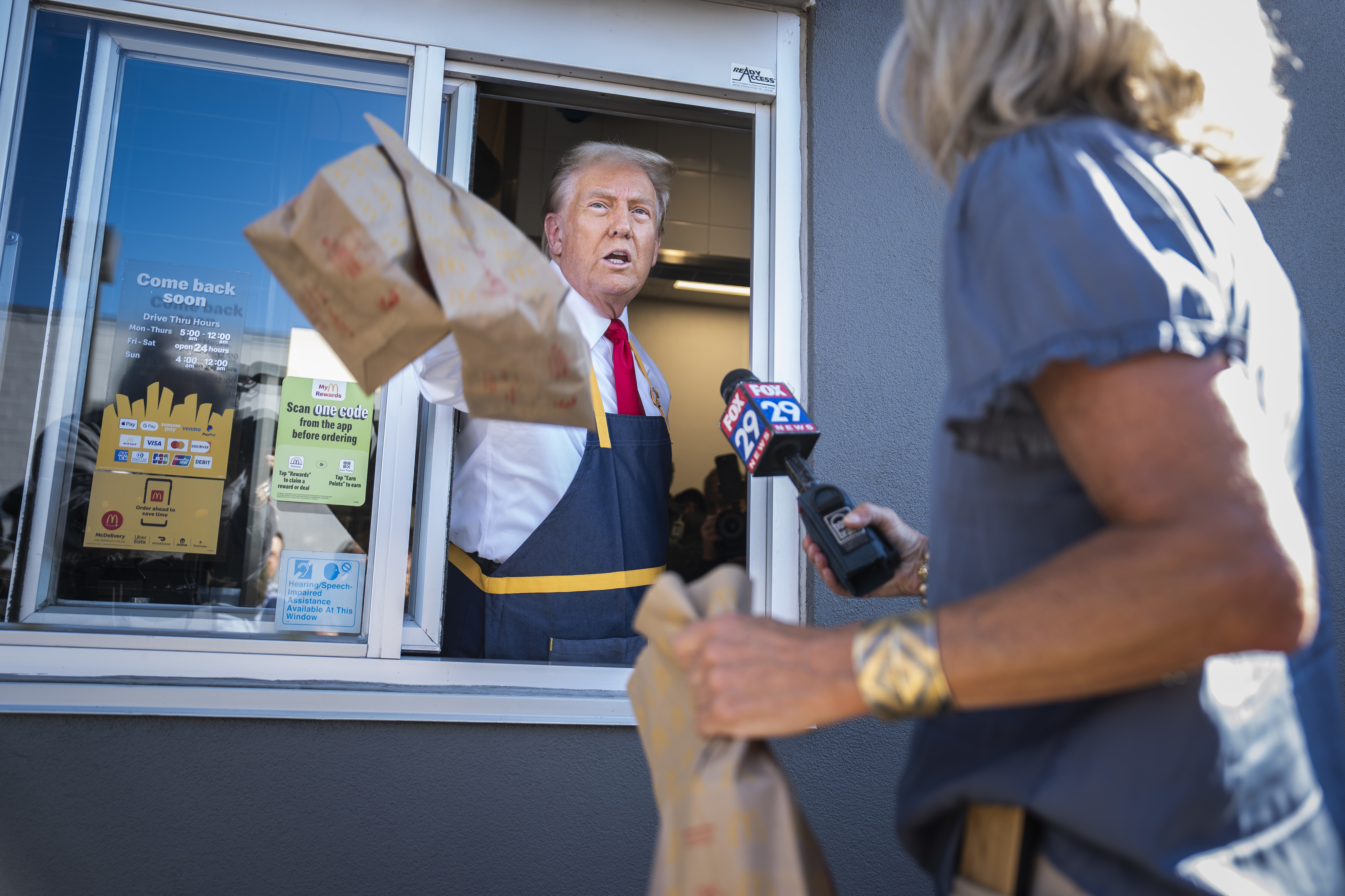 donald trump in a fast-food drive-thru hands bags out the window to a customer with a microphone