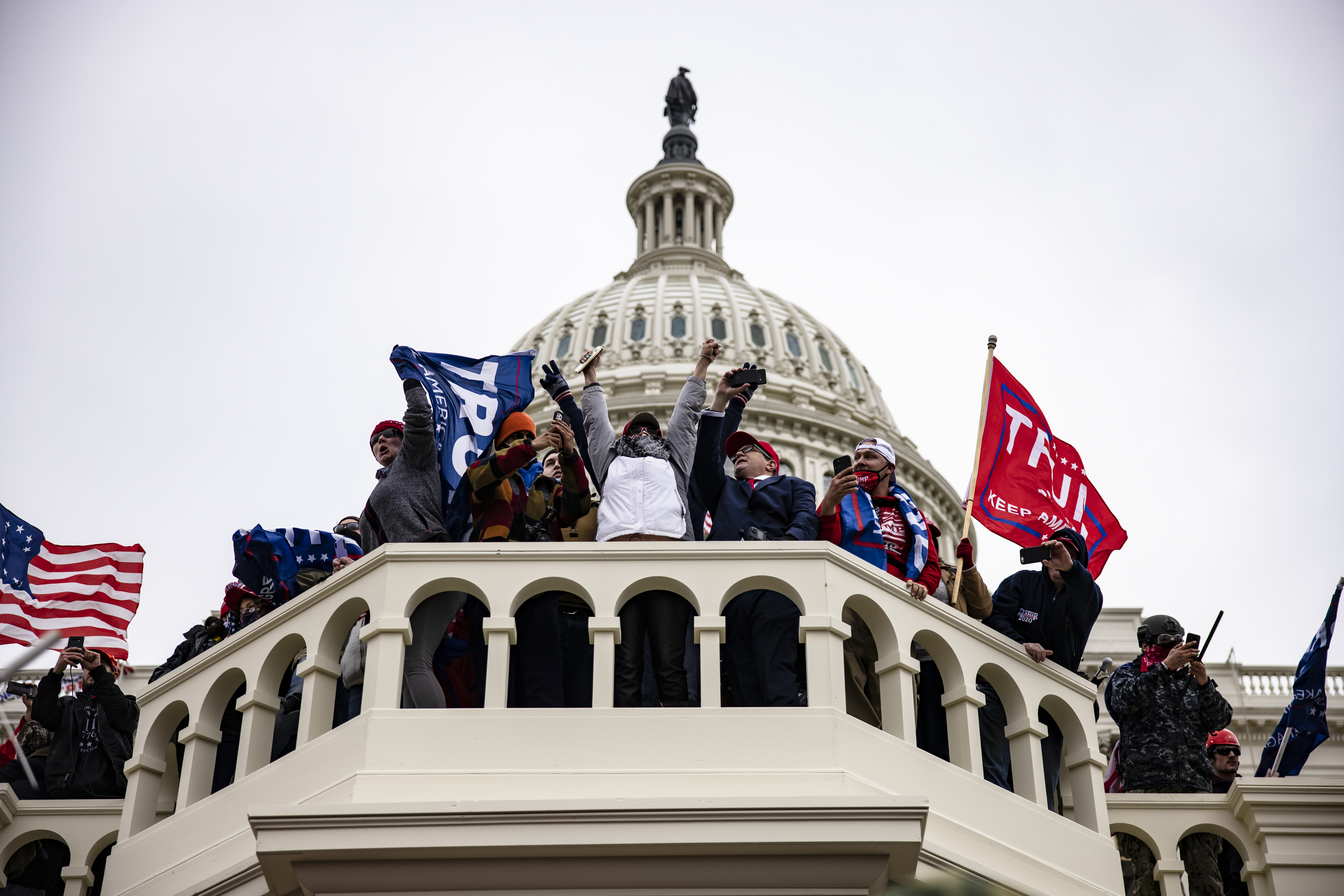 Protesters on U.S. Capitol balcony holding flags and wearing winter clothing, with the dome in the background