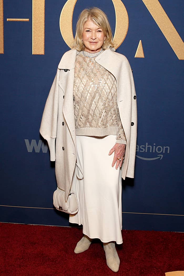 I don’t know who this is, but they're on a red carpet wearing a beige coat with a patterned sweater and white skirt