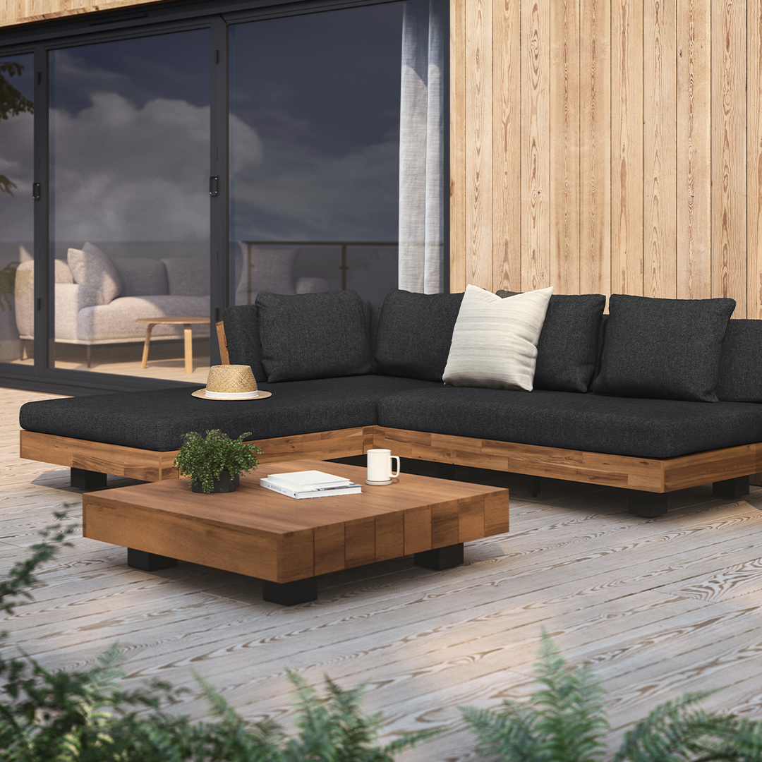 Outdoor wooden patio set with a dark cushioned L-shaped sofa, low square coffee table, and potted plants, set on a deck with glass doors in the background