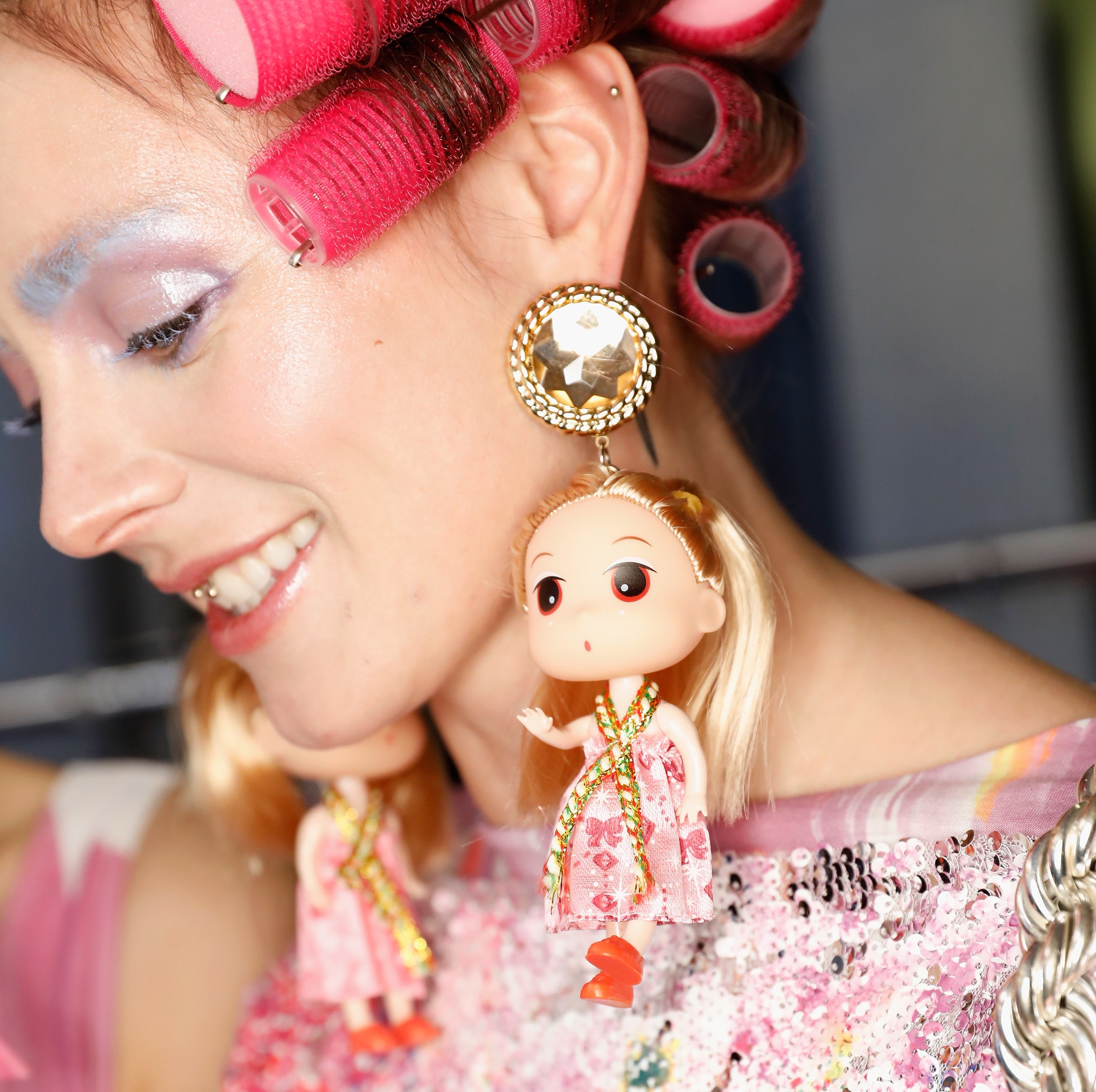 Person with hair curlers, wearing large doll-themed earrings and a sequin-patterned outfit, smiling